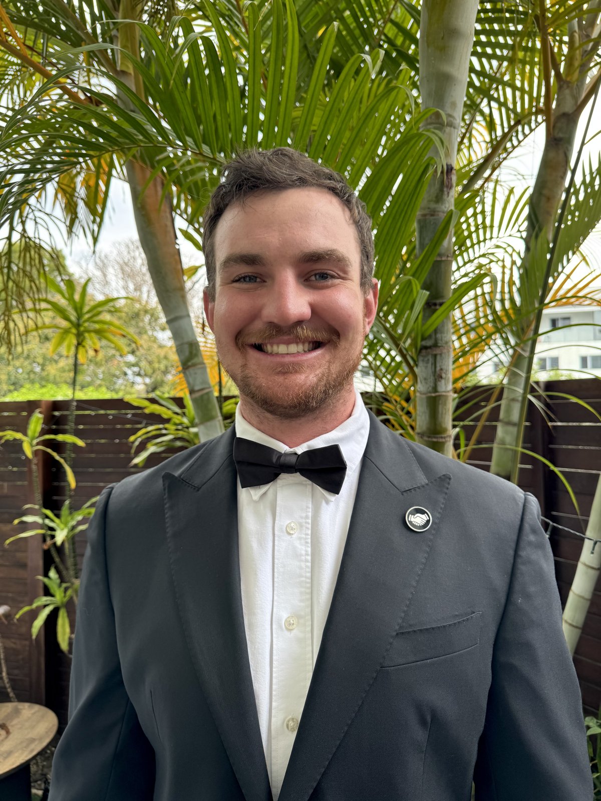 A man in a black suit and bow tie wearing a Mates pin on his lapel